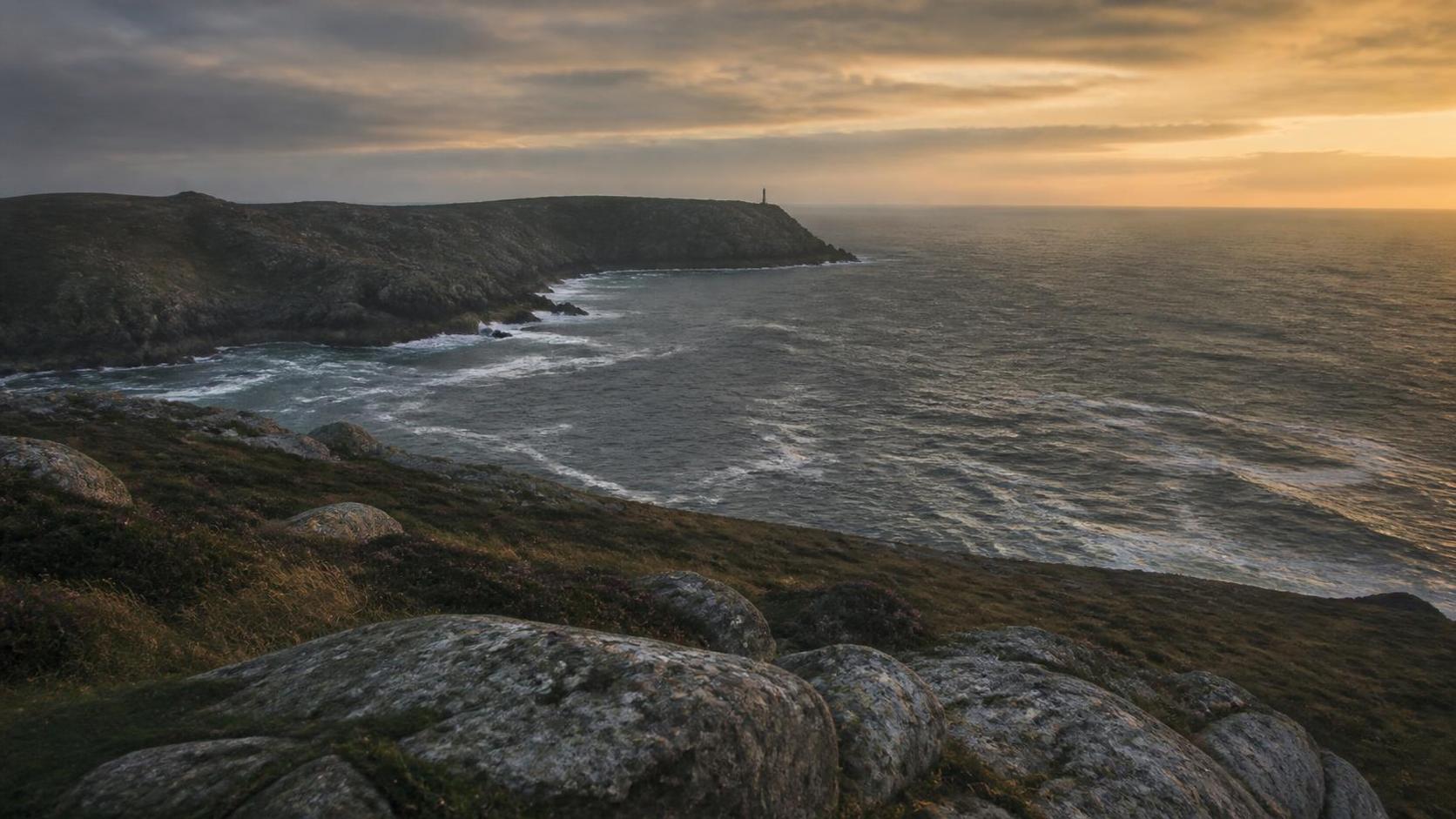 A rocky headland on the Galician coast at dusk, heather and granite in the foreground, a distant lighthouse on the cliff and the Atlantic breaking below a gold horizon.