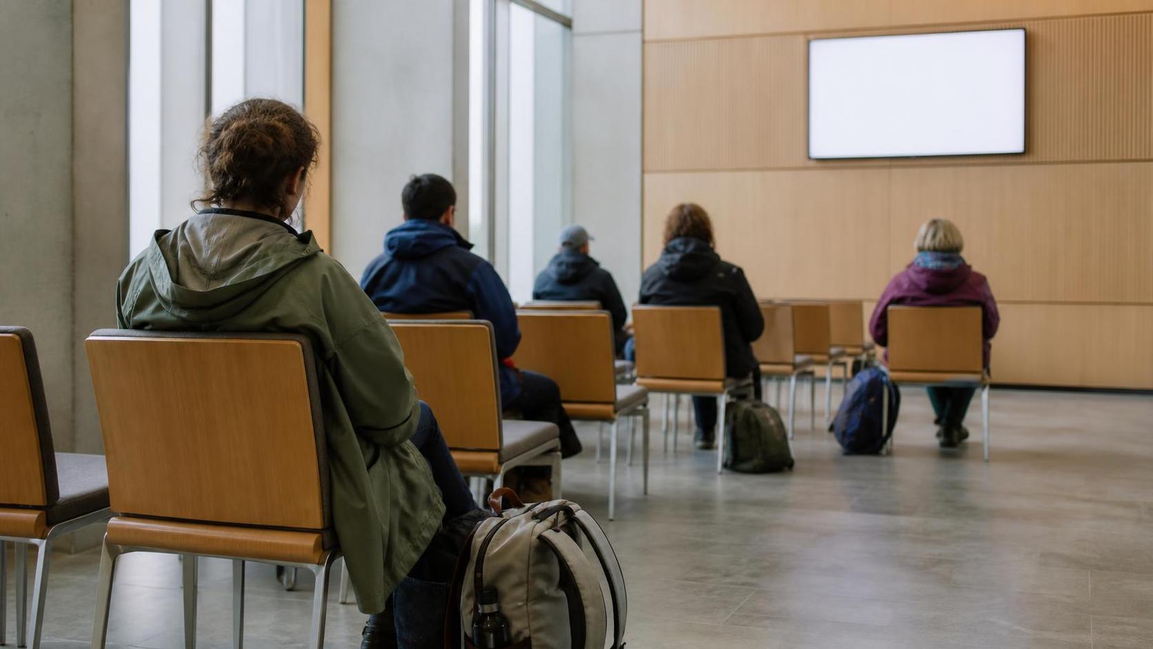 Travellers in rain jackets seated from behind on rows of chairs in a plain waiting room, small daypacks at their feet, facing a blank queue display on the wall.