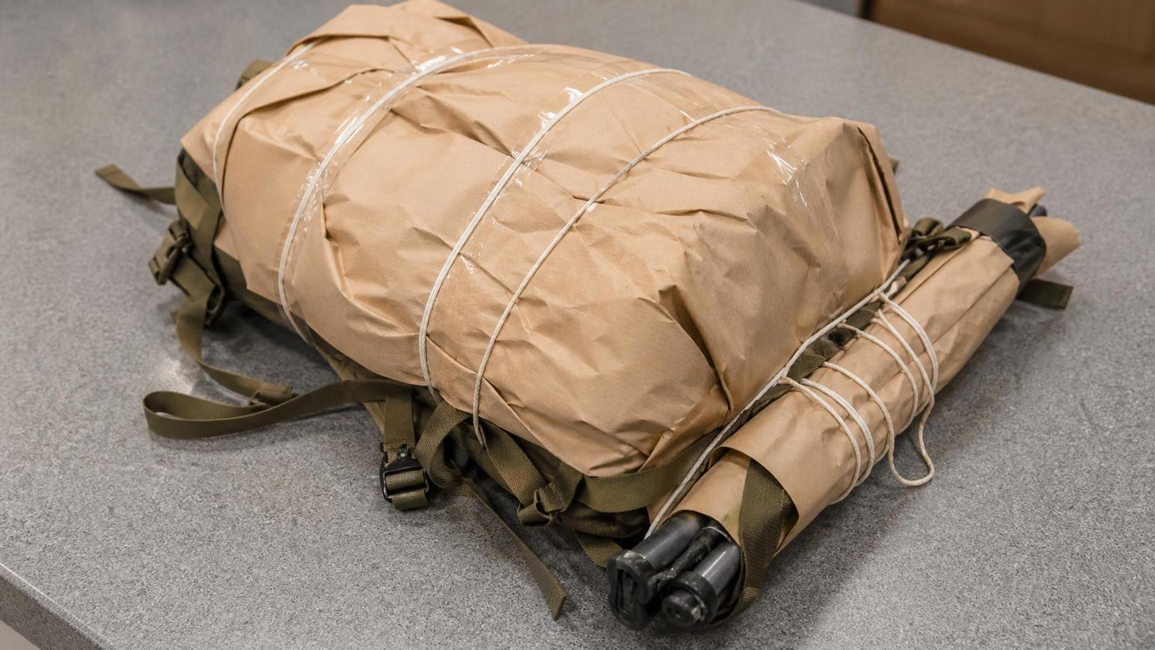 A hiking backpack wrapped in brown parcel paper and tied with string on a post office counter, a bundle of trekking poles wrapped alongside it.