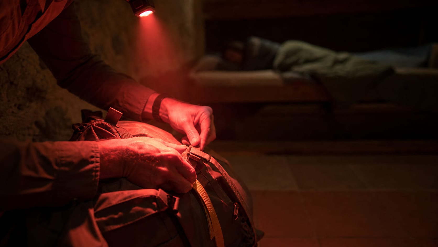 Close view of a pilgrim's hands closing a backpack before dawn in a dark dorm, lit only by the red beam of a head torch, a sleeping bunkmate soft-focused behind.