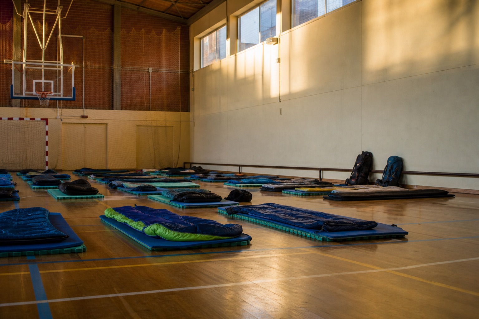 A small Spanish village polideportivo set up as pilgrim overflow accommodation, with foam mats and sleeping bags arranged on a wooden gym floor in soft evening light.