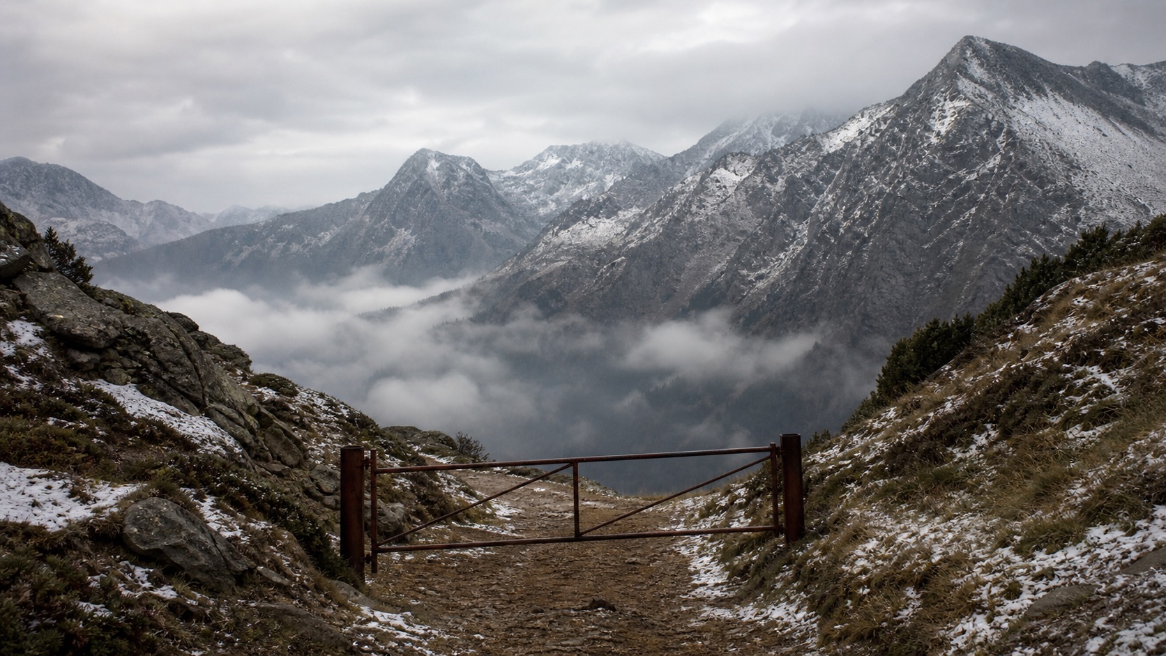A snow-dusted Pyrenees ridge in late autumn with a simple rusted metal gate across a trail, mist hanging in the valley below.