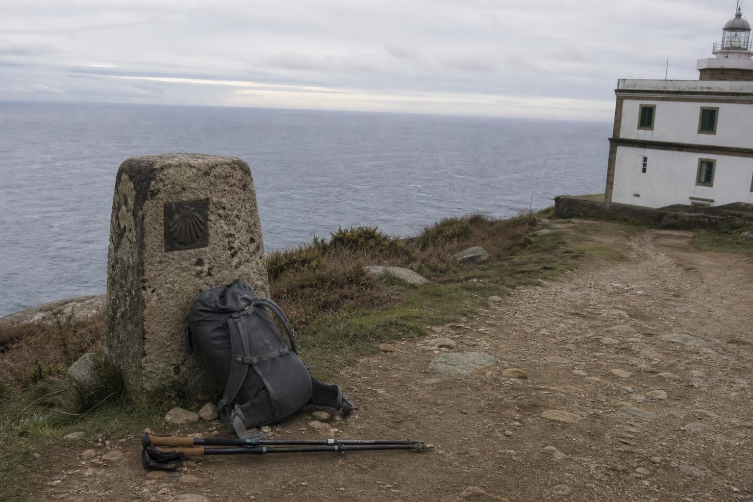 Ein verwitterter Wegstein aus Granit mit einer Jakobsmuschel aus Bronze am Schotterweg am Kap Fisterra, ein grauer Rucksack lehnt daran, Wanderstöcke liegen am Boden, rechts am Bildrand der Leuchtturm, dahinter der flache graue Atlantik.