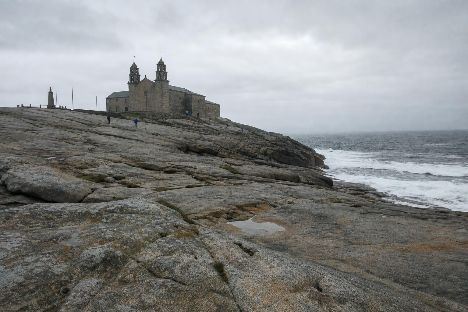 Die graue Granitkirche Virxe da Barca in Muxía mit ihren zwei Glockentürmen über breiten, flachen Granitplatten am Meer, flacher bedeckter Himmel, rechts bricht die Brandung an den Felsen.