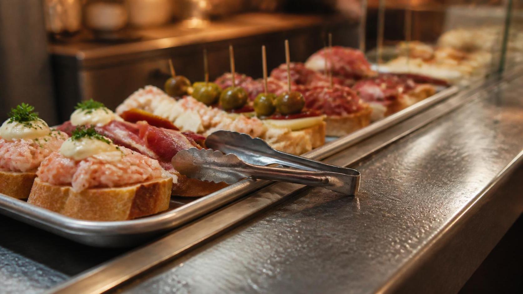 Ein Edelstahltablett mit gemischten Pintxos auf Brotscheiben auf einer spanischen Bartheke, quer darüber eine einzelne gemeinsame Metallzange.
