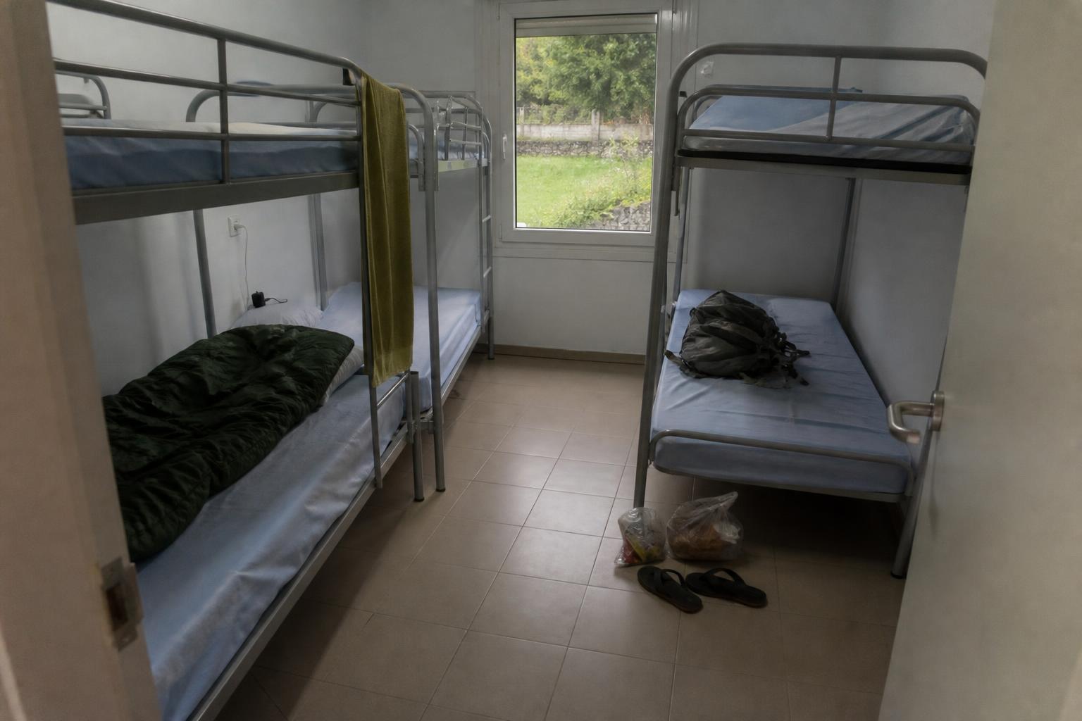 Una pequeña habitación de albergue público con literas metálicas grises, solo dos ocupadas, un saco de dormir verde en una y una mochila en otra, bolsas de plástico con comida y chanclas en el suelo de baldosas.