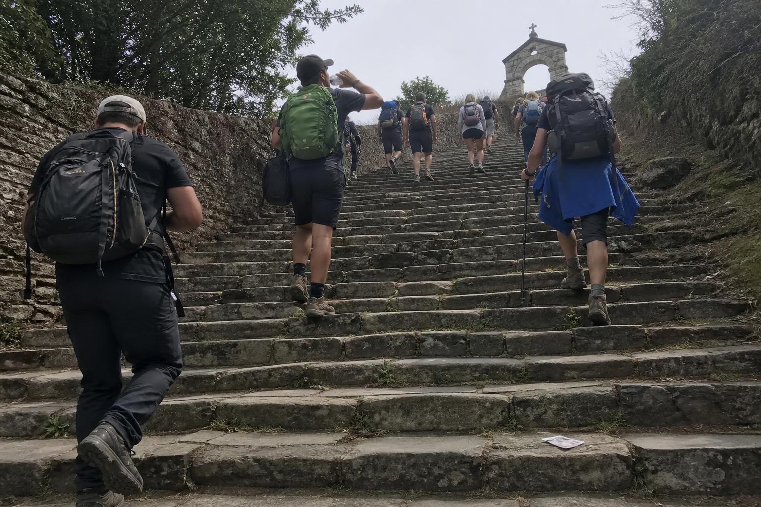Una docena de caminantes con mochila subiendo una amplia escalinata de granito hacia el pequeño arco de piedra de una capilla en Portomarín, todos vistos de espaldas.