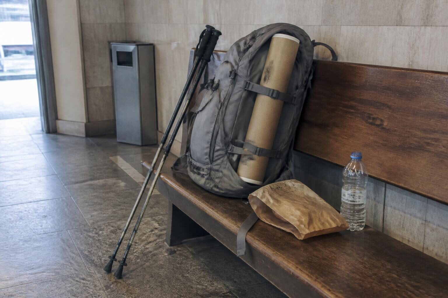 A dusty grey hiking backpack on a worn wooden bench with a plain brown cardboard document tube tucked upright under its side straps, trekking poles leaning beside it.